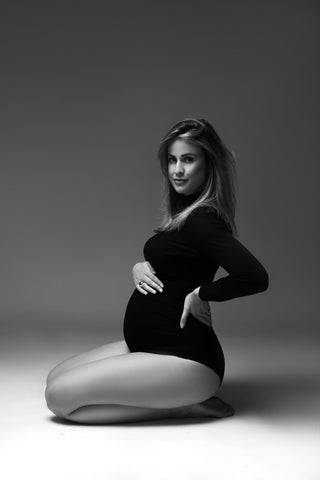 pregnant model poses in a studio wearing a black bodysuit with long sleeves and a turtleneck neckline. 