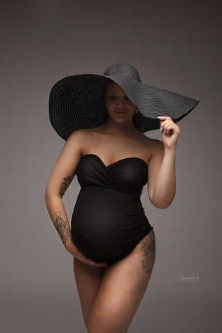 pregnant model poses wearing a strapless black bodysuit and a big black hat.