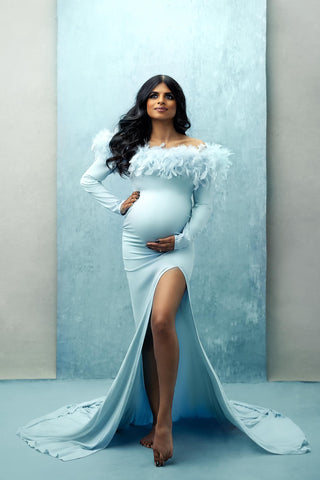 modèle aux cheveux foncés pose dans un studio sur un fond bleu clair vêtu d'une robe bleu clair longue et serrée à manches longues et d'un haut recouvert de plumes bleu clair.