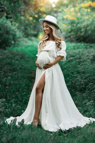 mannequin enceinte blonde pose à l'extérieur vêtue d'un ensemble bohème blanc composé d'une jupe et d'un haut. elle a un chapeau blanc pour correspondre au style.