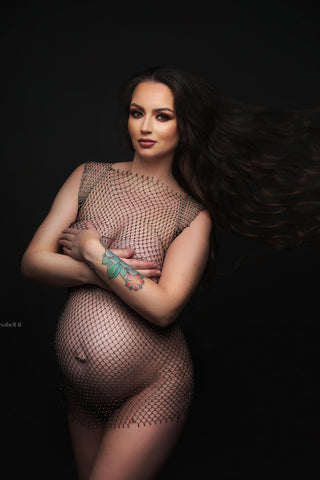 brunette pregnant model poses against a black background wearing a see through piece of clothing. she covers her chest and stares to the camera.