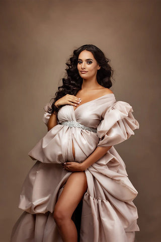 Brunette model poses in a studio wearing a taffeta long dress in sand color. the dress has a lot of ruffles details and a sash to match the style.