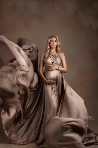 blond pregnant model poses in a brown studio wearing a dusty pink silk dress. there is a lot of flowing fabric during the shoot.