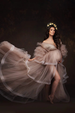Brunette model poses in a studio wearing a long dress with a lot of tulle layers. the dress is in sand color and has a bodysuit underneath and several tulle ruffles all over the dress.