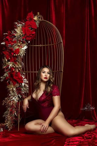 modèle blond foncé pose dans un studio, assis à côté d'une chaise en métal qui ressemble à une cage avec plusieurs détails rouges. elle porte un body rouge lors d'une séance boudoir.