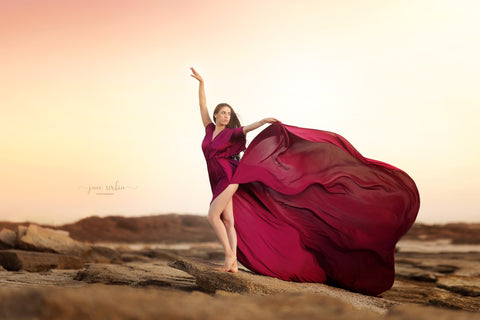 modèle pose au bord de l'eau portant une robe soyeuse de couleur cerise avec une traîne extrêmement longue.