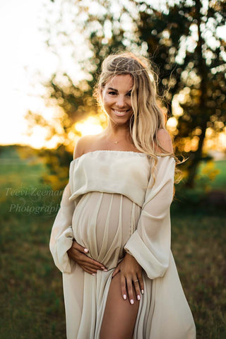 blond pregnant model poses outdoors during the golden hour wearing a chiffon set in sand color. the top features long sleeves and an off shoulder neckline. there is a split on the skirt where one of her legs can be seen.