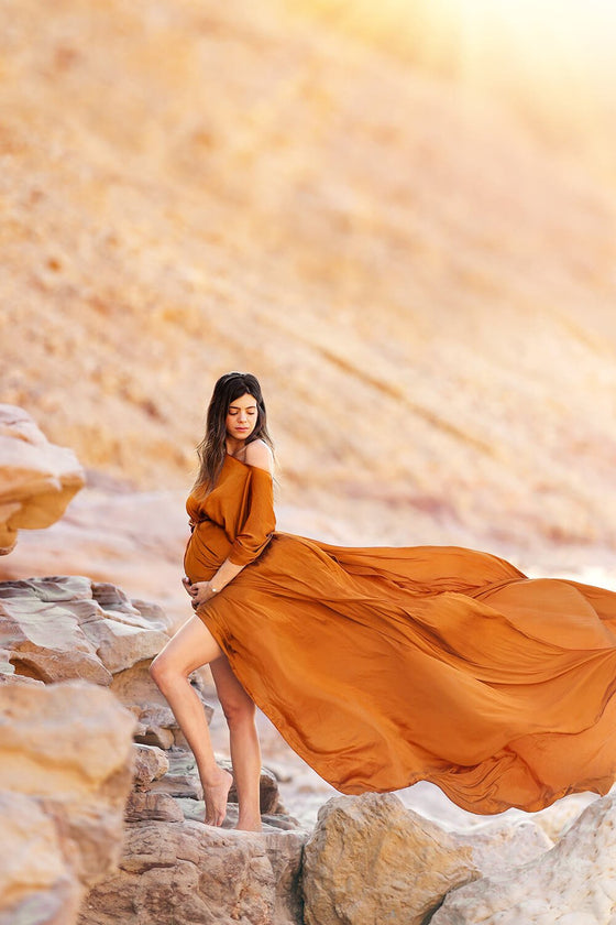 pregnant brunette model is standing in a place full with big stones. she is looking to the floor and posing holding her baby bump. she is wearing a silky cognac very long maternity cape. 
