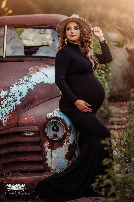 Pregnant model poses outside next to an old car wearing a long black dress completely tight, with a turtle neck neckline and long sleeves. She also wears a hat which she is touching with one hand while the other is holding her bump. Model looks away and wears jewelry. 