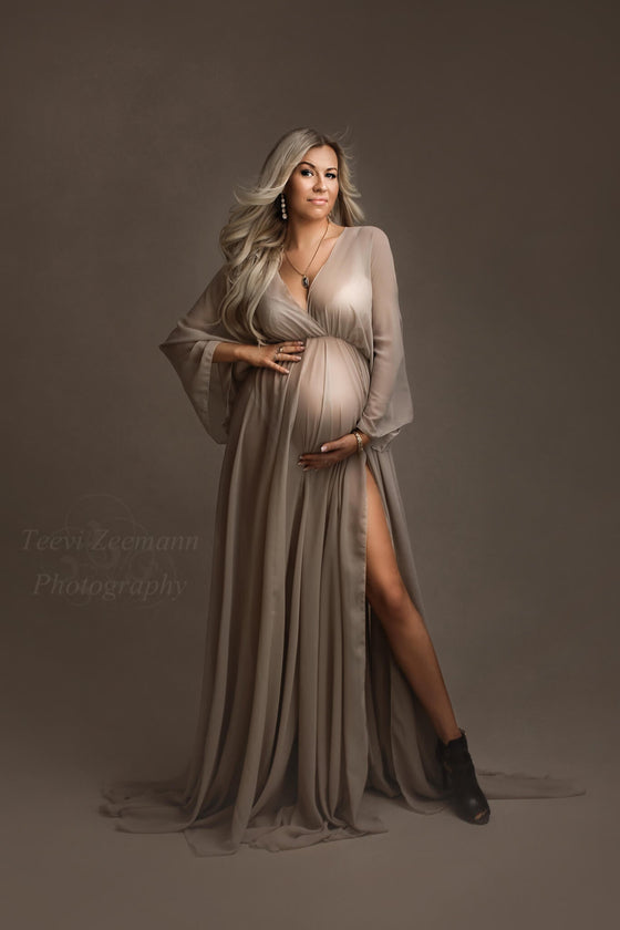 Chiffon long dress in khaki is being used by a blond pregnant model. She poses during a maternity photoshoot in a studio. The dress features a low-v cut neckline and kaftan sleeves. The model faces the camera and wears boots and jewelry to complete the look. The dress has a split on the side where you can see one of her legs through. 