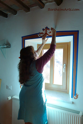 The artist Kelly removes the painter's tape on the first window after the acrylic paint has dried while painting a different window.