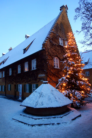 a tall lit Christmas tree matches the shape of the pointed roof in Augsburg Germany the well is also covered with snow