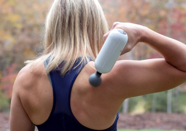 Woman using massage gun on muscles around shoulder blades.