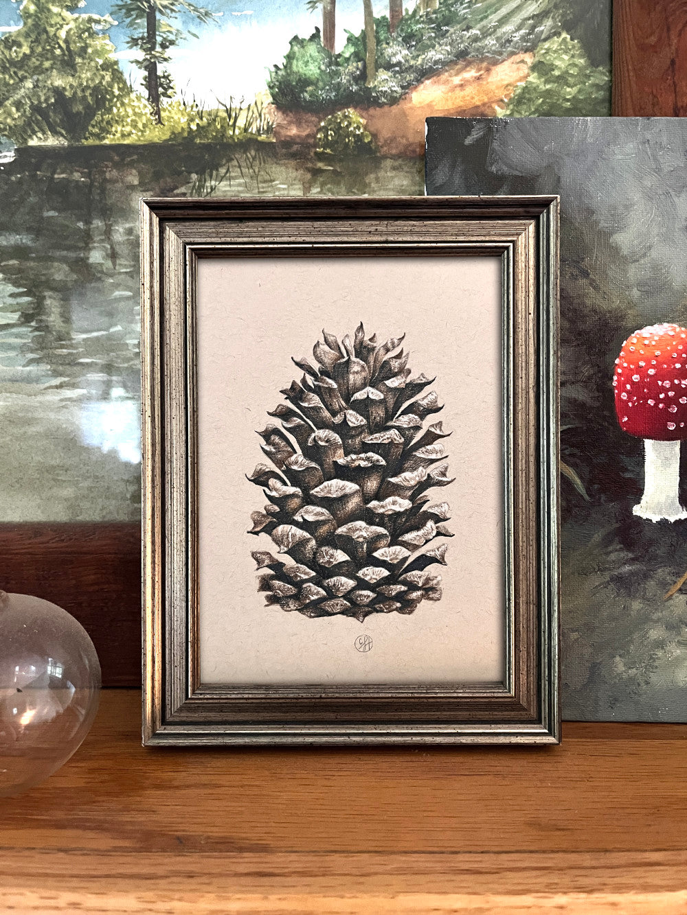 pinecone artwork, framed, on a mantle with other nature art surrounding it.