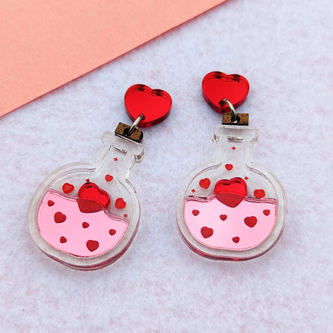 Love potion bottle earrings made with clear acrylic with shiny pink for the liquid and red for the hearts. A wooden cork is at the top and they hang from shiny red heart stud posts.