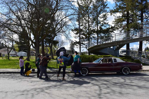 Wide shot of outdoor photoshoot. Andrea is in front of a vintage maroon Cadillac. The photographer and multiple helpers are surrounding the car.