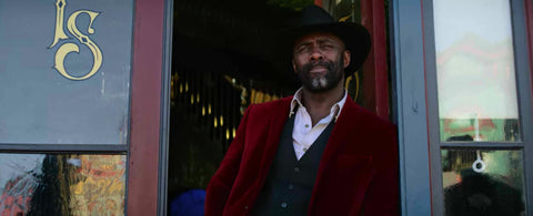 Rufus Black, played by Idris Elba, leans in a doorway with a deep red velvet jacket on.