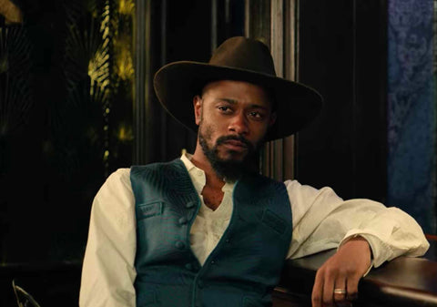 Cherokee Bill, played by LaKeith Stanfield, in a white shirt with blue vest. He is leaning against a bar on one elbow.