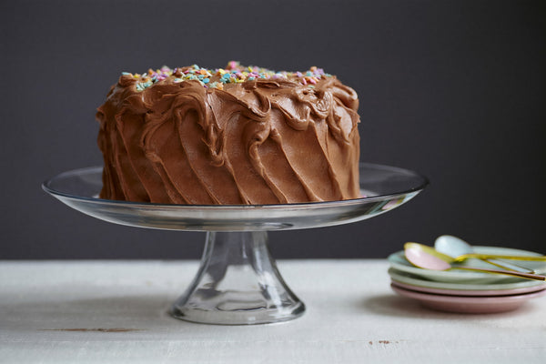 Chocolate cake on a stand with stacked plates and spoons in the background