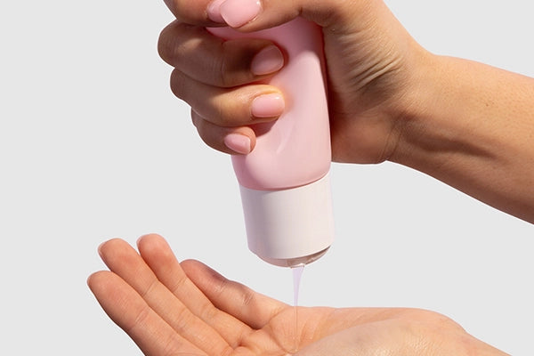 Hand squeezing pink tube of lubricant onto other hand against white background