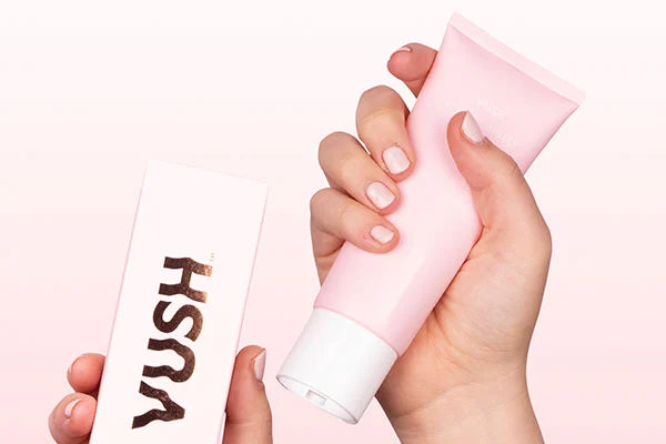Hand holding VUSH pink cardboard box while another hand holds VUSH Intimate Gel in pink tube with white lid against light pink background