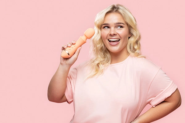 Blonde woman holding VUSH orange Majesty 2 wand vibrator in right hand and smiling against light pink background