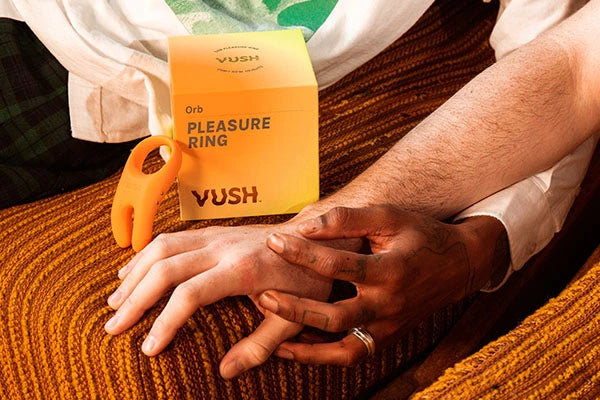 Two hands holding on orange couch next to orange box that reads "PLEASURE RING | VUSH" and orange vibrating cock ring