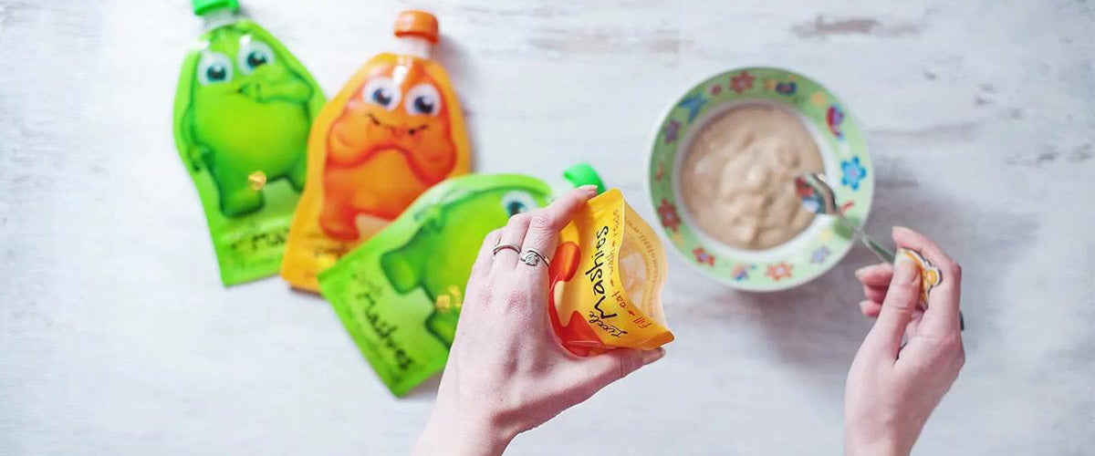 Reusable food pouches being filled with baby food. A woman is holding open a reusable food pouch in one hand, whilst spooning baby food from a bowl and into the pouch with the other hand. Next to the bowl lies two more reusable food pouches ready to be filled.