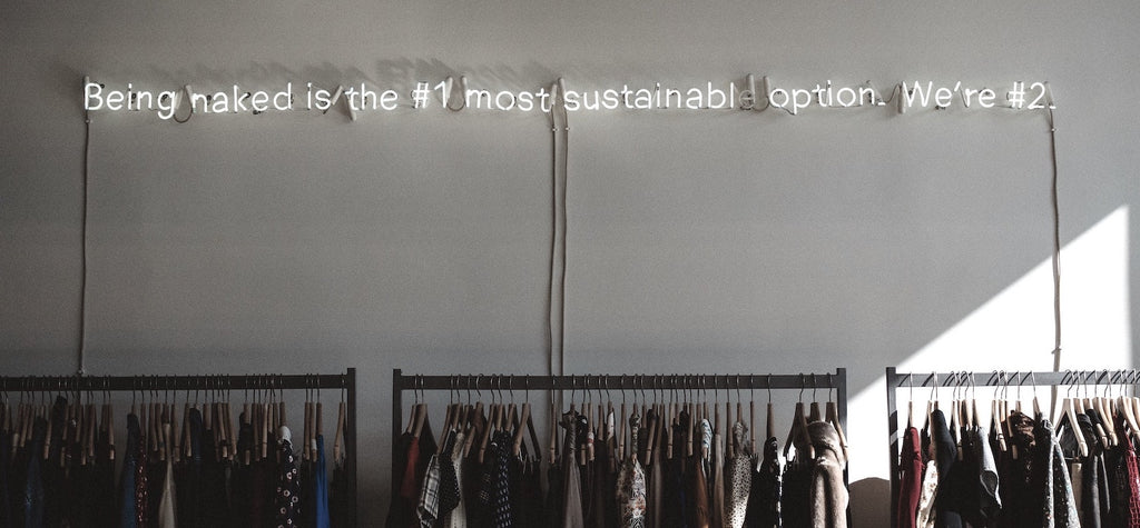 racks of clothing attached to a white wall with a neon sign above that says "being naked is the number one sustainable option. We are number two."