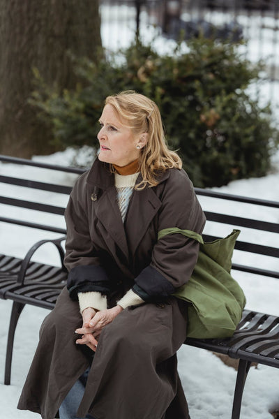 elderly woman sitting on a bench with a green tote bag in winter