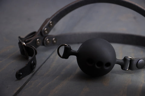 A closeup of a leather head harness ball gag, showing the snap in and snap out functionality on the ball gag, and how the silicone ballgag has three holes for breathing. 