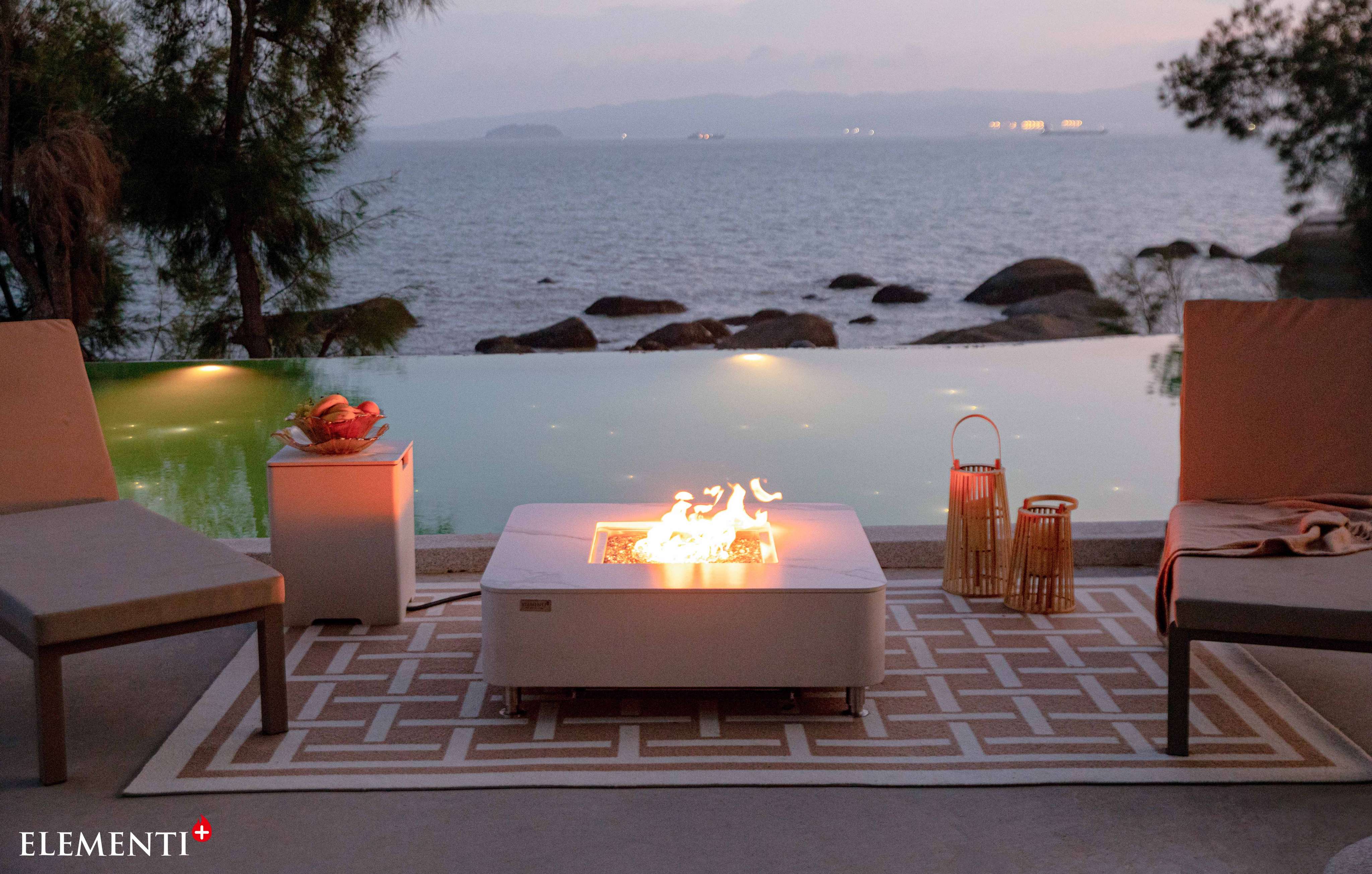 The Elementi Plus Annecy Porcelain Top Fire Table is positioned on a patio beside the water. 
