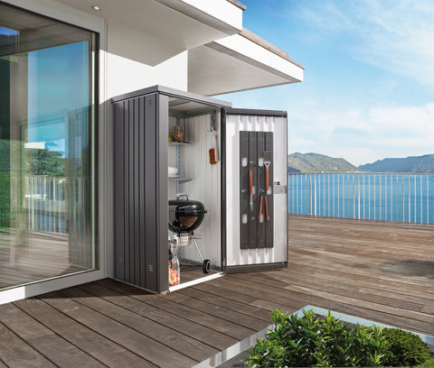 Biohort Equipment Locker 90 in dark gray with the door open, showcasing storage space with shelves containing garden tools and a barbecue grill, situated on a wooden deck with a scenic mountain and lake backdrop.
