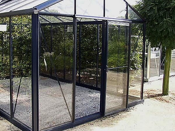 Janssens Royal Victorian Greenhouse with stylish black frame and glass panels, furnished as an outdoor living space with a fire pit and wicker furniture.