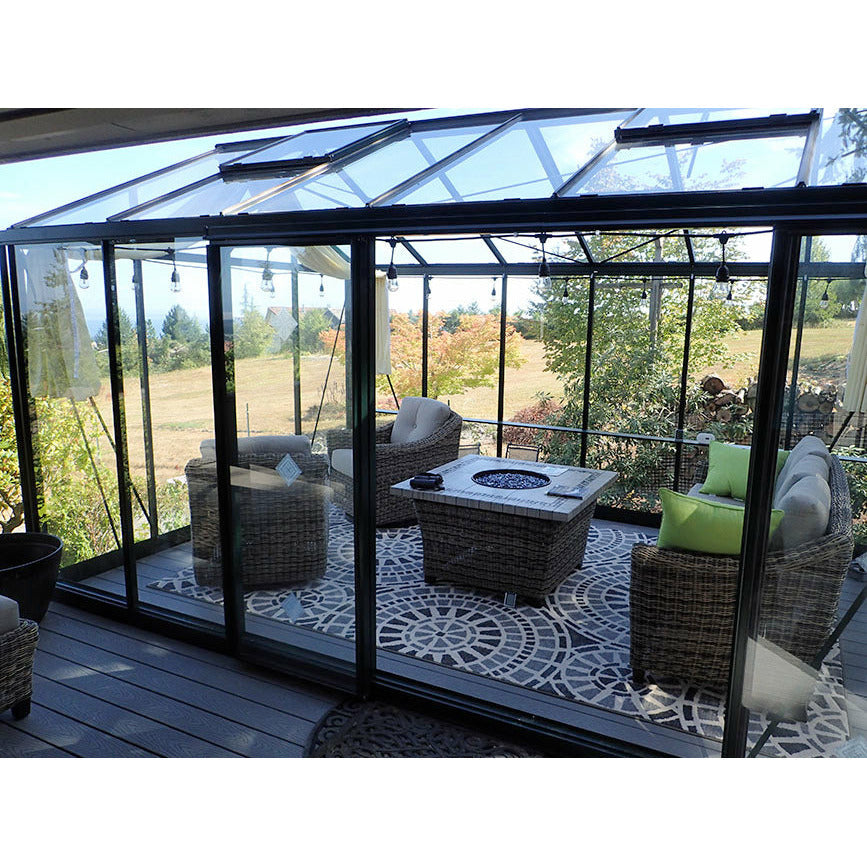 Janssens Royal Victorian Greenhouse with stylish black frame and glass panels, furnished as an outdoor living space with a fire pit and wicker furniture.