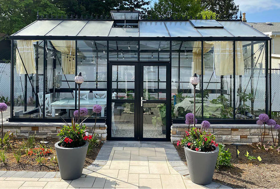 Royal Victorian greenhouse with hinged double doors, featuring a landscaped garden and patio, surrounded by lush plants and flowers.