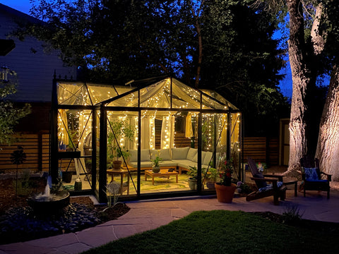 Exaco Janssens Royal Victorian Orangerie Greenhouse illuminated by warm lights at twilight, creating a cozy outdoor retreat with seating and vibrant plant life.