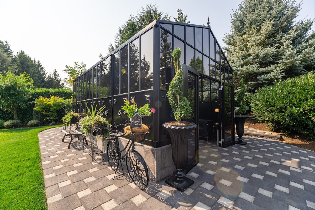 The Exaco Janssens Retro Royal Victorian VI Greenhouse standing on a patterned patio, enhanced by potted plants and vintage-style garden furniture.