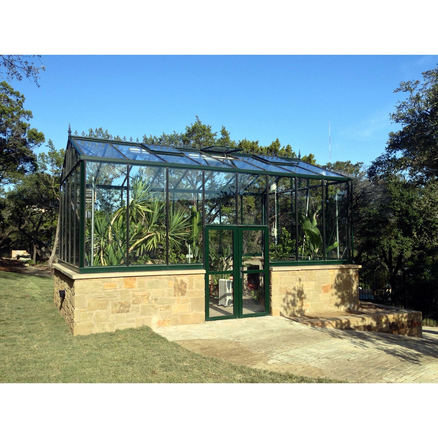 Exaco Janssens Royal Victorian VI 46 Greenhouse perched on an elegant stone foundation, surrounded by towering greenery and clear blue skies.