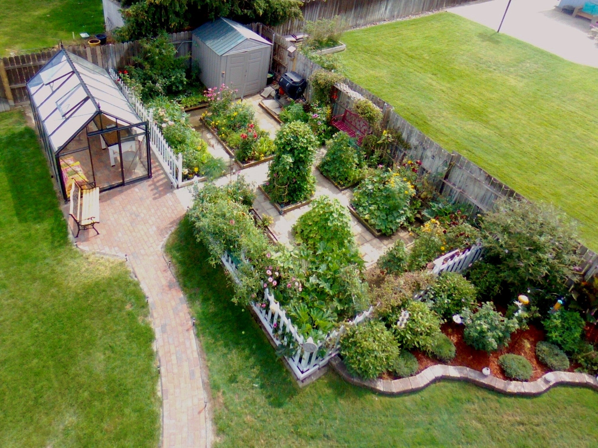 Aerial view of an Exaco Junior Victorian styled greenhouse situated in a meticulously landscaped garden with raised flower beds, winding brick pathways, and lush greenery.