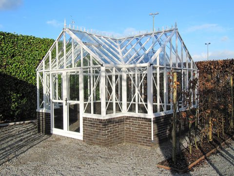 A standalone Victorian greenhouse with a gable roof, made of clear glass and white aluminum frames on a brick base. It's placed in an open space with a trimmed hedge in the background.
