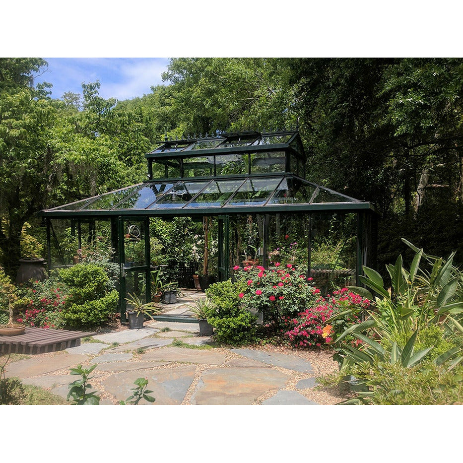 A 15x20 ft Victorian cathedral greenhouse surrounded by lush garden, with a distinctive large cupola and green framework.