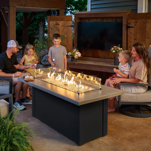 A family enjoys an outdoor gathering around the Fire Pit Table, under a pergola with string lights.