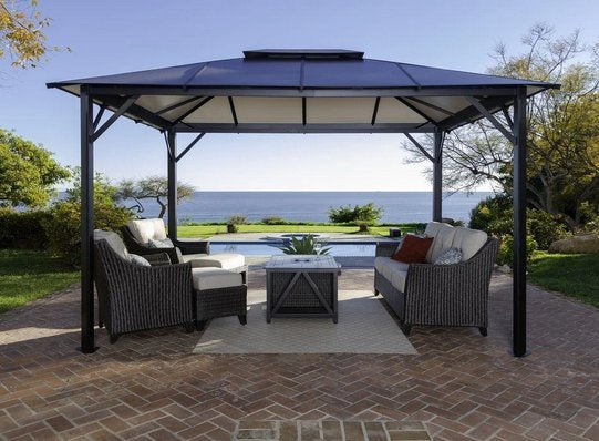 Paragon Outdoor Durham Hard Top Gazebo set on a brick patio with comfortable outdoor seating, overlooking a tranquil seaside view.