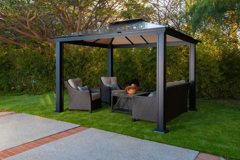 Paragon Outdoor Santa Monica Hard Top Gazebo set in a lush garden, furnished with a cozy wicker love seat and chairs with plush cushions.