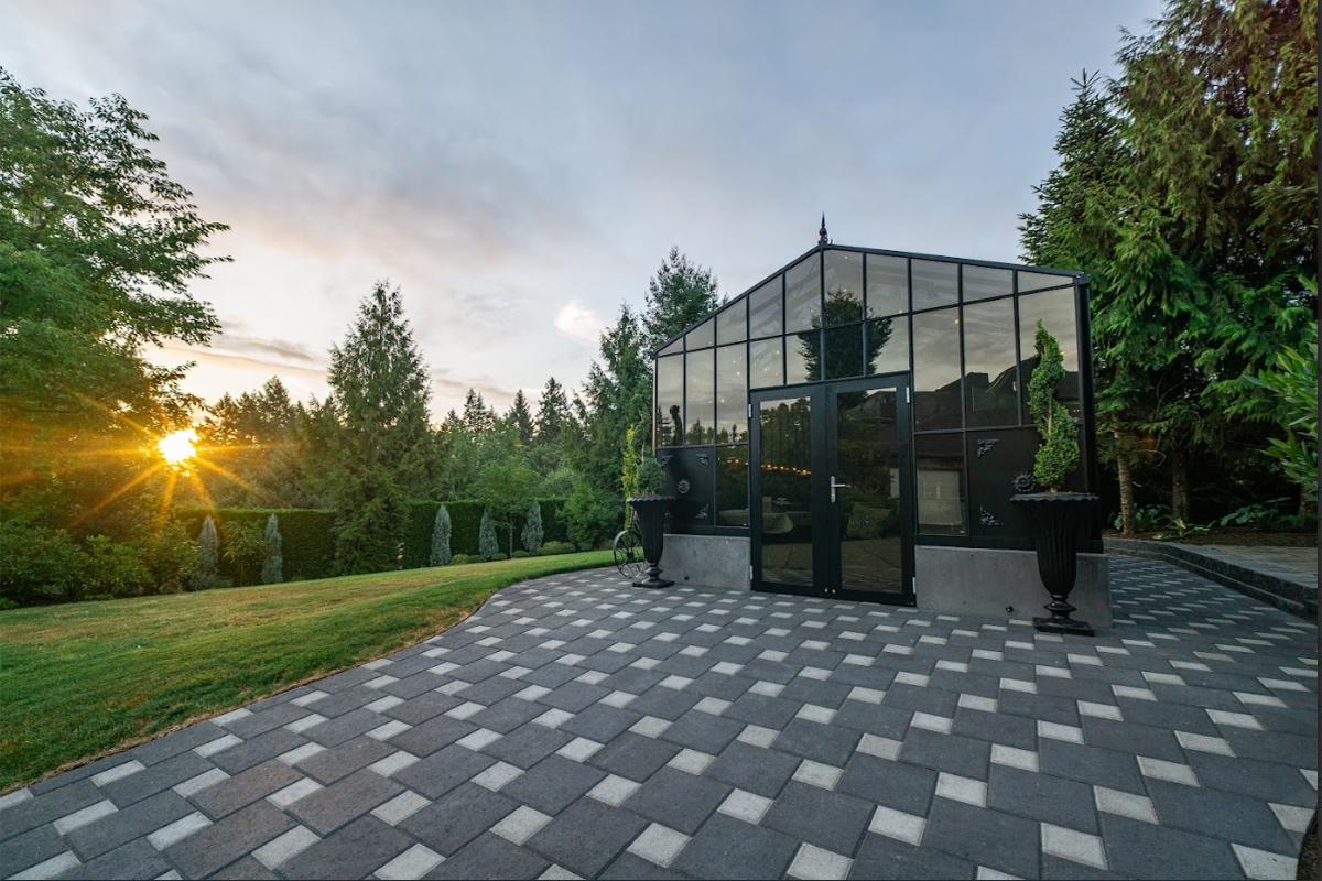 The Exaco Janssens Retro Royal Victorian VI Greenhouse bathed in the golden hues of sunset, with a checkered patio and lush greenery surrounding it.