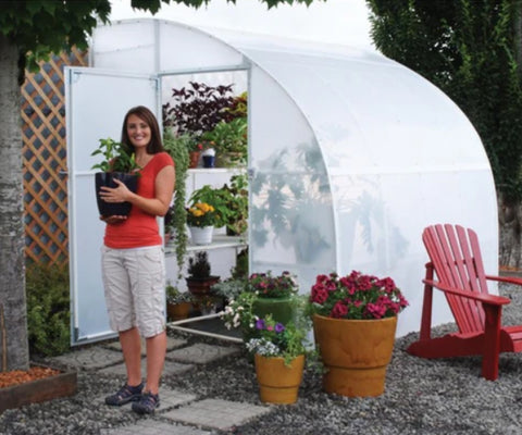 A woman standing outside the Solexx Harvester Greenhouse Kit holding up a plant. There are plants displayed on the shelves inside.