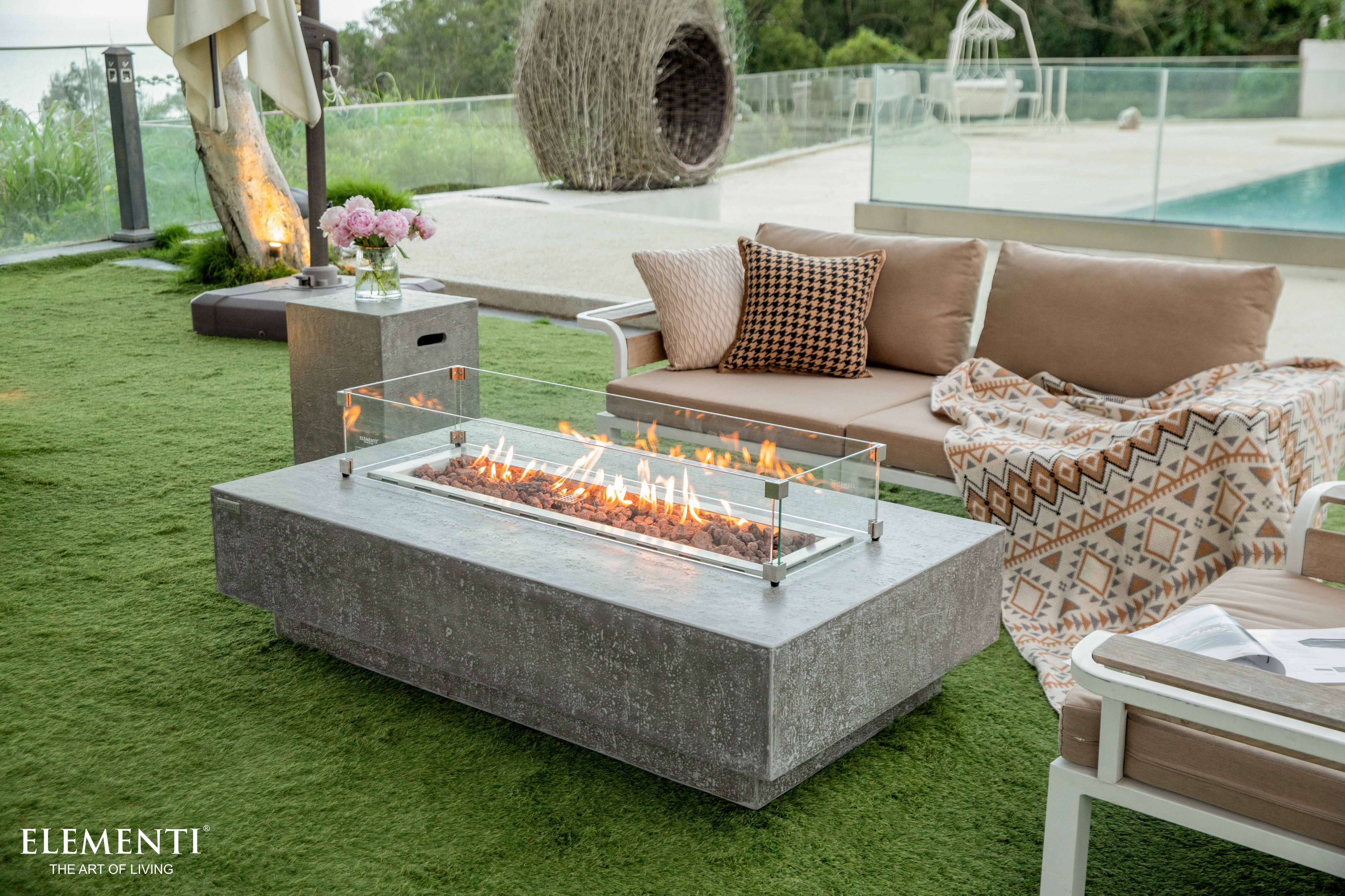 The Elementi Hampton Fire Table, featuring a flame guard, rests on grass adjacent to a serene pool, enhancing outdoor elegance