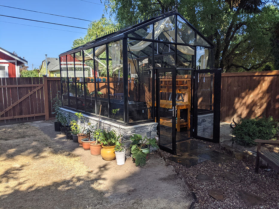 A Royal Victorian VI23 greenhouse with green frame, open roof vents, and white shade cloths in a lush garden.