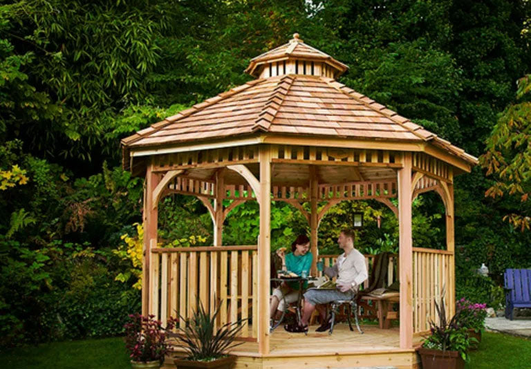 a garden space with the Outdoor Living Today Bayside Gazebo with a couple sitting inside it and with trees in the background